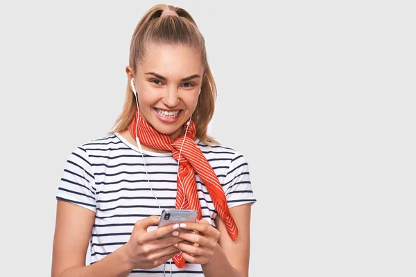 Jovem europeia feliz com penteado de rabo de cavalo, vestindo camiseta listrada, elegante lenço vermelho no pescoço, olhando para a câmera ouvindo suas músicas favoritas através de fones de ouvido brancos usando smartphone — Fotografia de Stock