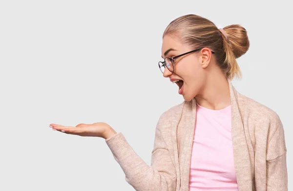 Zszokowana szczęśliwa Blondynka Ładna młoda kobieta wygląda przyjemnie do dłoni, pokazując coś w ręku, nosząc okrągłe przezroczyste okulary i casual beżowy i różowy bluzki z pustą przestrzenią kopiowania — Zdjęcie stockowe