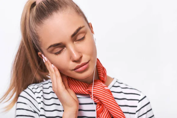 Retrato de cerca de una hermosa joven con una camiseta a rayas, elegante bufanda roja en el cuello, relajándose con los ojos cerrados escuchando sus canciones favoritas a través de auriculares blancos usando un teléfono inteligente —  Fotos de Stock