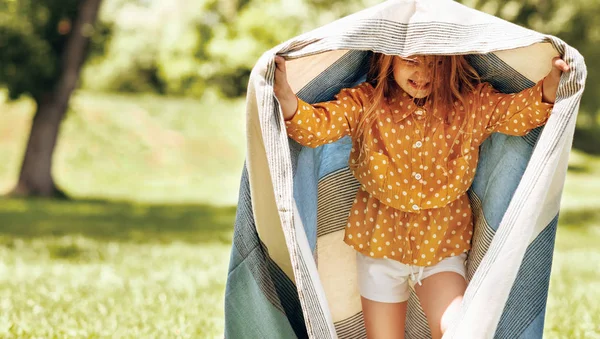 Imagem ao ar livre da menina loira feliz brincando com o cobertor, sorrindo amplamente, aproveitando o verão. Criança adorável se divertindo sob o cobertor durante as férias com sua família no parque . — Fotografia de Stock