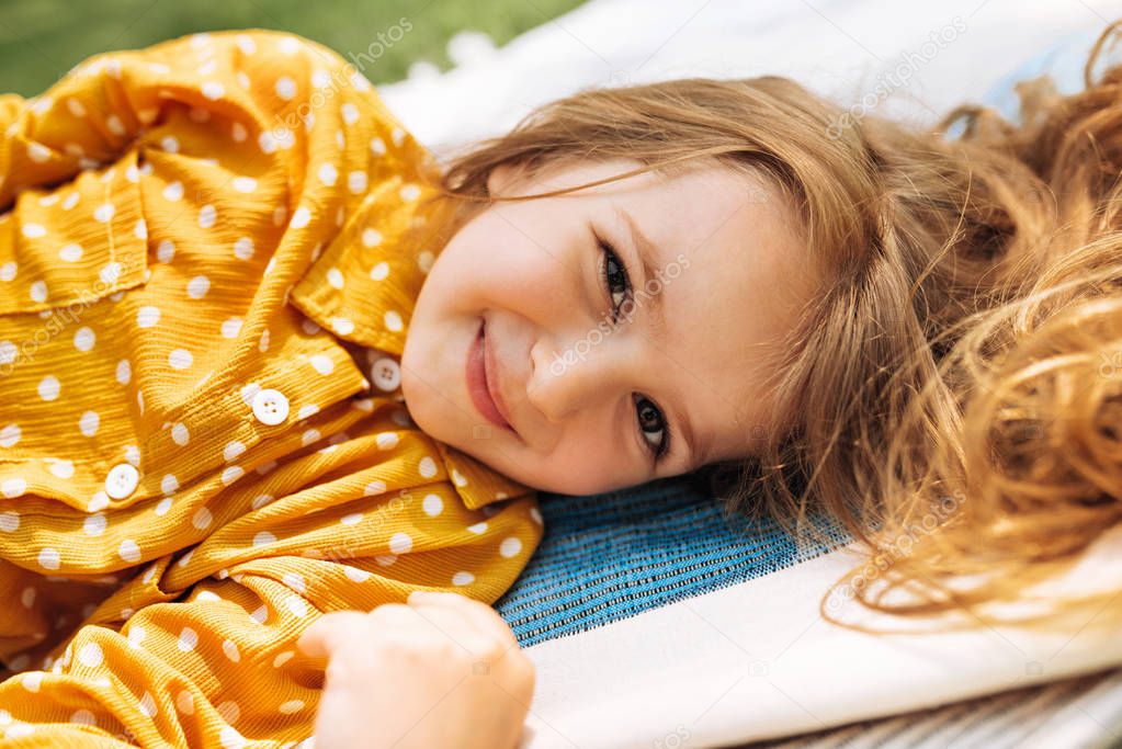 Closeup portrait of happy little girl smiling and lying on the blanket at grass, enjoying summertime outdoors. Adorable child having fun and relaxing during picnic with her family in the park.