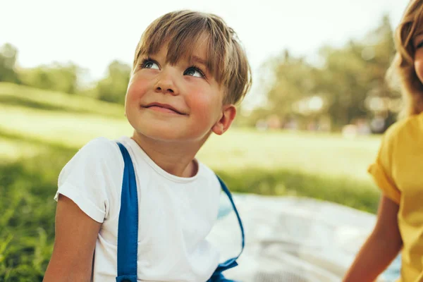 Улыбающийся мальчик, сидящий на одеяле на фоне природы. Счастливый ребенок наслаждается летом со своей сестрой в парке. Красивый ребенок улыбается и веселится на солнечном свете на открытом воздухе. Детство — стоковое фото
