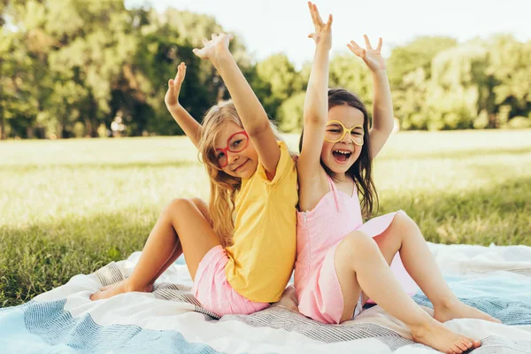 잔디에 담요에 앉아 사랑스러운 아이들. 공원에서 여름을 즐기고, 종이 안경을 입고 행복 두 어린 소녀. 두 자매는 야외에서 미소를 지으며 햇빛을 즐기고 있습니다. 어린 시절 — 스톡 사진