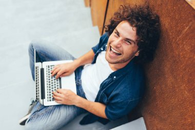 Şehir caddesinde ücretsiz kablosuz bağlı arkadaşlarınızla online sohbet için dizüstü bilgisayar kullanarak kıvırcık saçlı mutlu adam üst görünümü. Yakışıklı erkek dizüstü bilgisayar ile beton merdivenlerde oturuyor. Yaşam tarzı