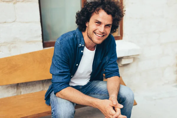 Imagem de jovem bonito inteligente com cabelo encaracolado, olhando para a câmera, sorrindo amplamente e sentado no banco na rua. Estudante do sexo masculino feliz relaxar ao ar livre. Pessoas e conceito de estilo de vida — Fotografia de Stock