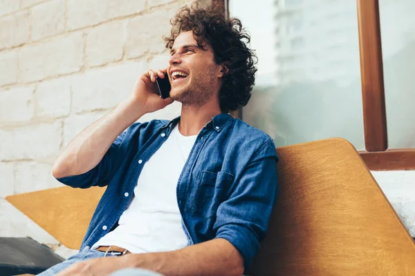 Jonge blanke man die buiten op de bank zit en praat met een mobiele telefoon. Gelukkige man met krullend haar die buiten aan het bellen is op zijn mobiele telefoon in de stad straat. Lifestyle en mensen — Stockfoto