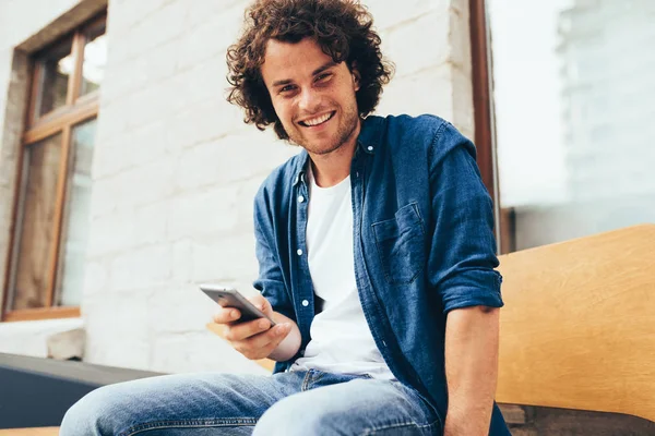 Smiling young man stitting outdoors texting on mobile phone. Happy male with curly hair using smart phone application for searching cafes near location in downtown in the city. Lifestyle and people — Stock Photo, Image