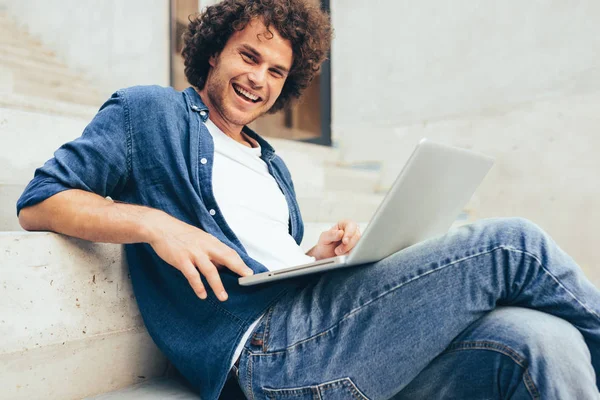 Kaukasische vrolijke man met krullend haar met behulp van laptop voor online chatten met vrienden, surfen op het Internet op de straat van de stad. Knappe man glimlacht breed, texting berichten op zijn laptop computer. — Stockfoto