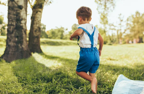 Immagine vista posteriore di bambino felice con indosso t-shirt bianca e pantaloncini blu che giocano sullo sfondo della natura. Bambino allegro che corre sull'erba verde nel parco. Il ragazzo si diverte all'aperto. Infanzia. . — Foto Stock