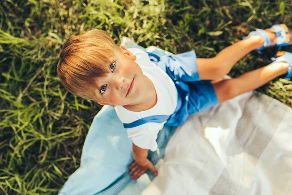 Над просмотром изображения симпатичного мальчика, сидящего на одеяле и смотрящего в камеру. Счастливый ребенок наслаждается летом в парке. Красивый ребенок улыбается и отдыхает на солнечном свете на открытом воздухе. Детство — стоковое фото