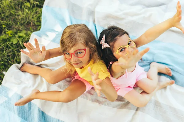 Über der Ansicht positiver Kinder mit Papierbrille mit erhobenen Händen, die auf der Decke sitzen. zwei kleine Mädchen genießen die Sommerzeit im Park. Freunde, die sich im Freien an der Sonne amüsieren. Kindheitskonzept — Stockfoto