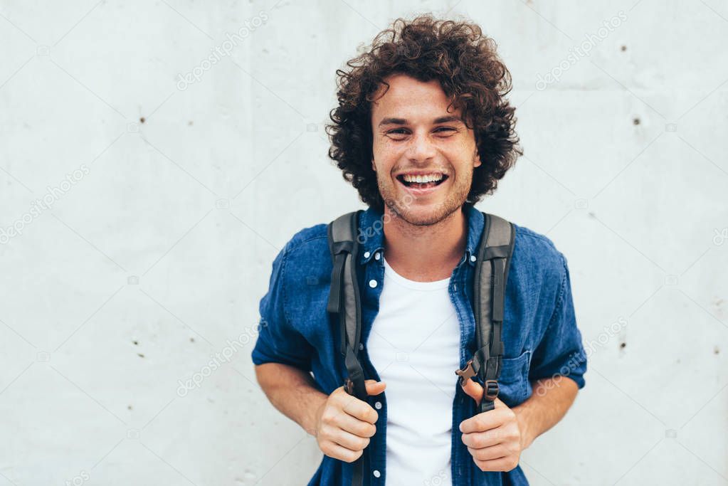 Outdoors horizontal portrait of young man with curly hair, smiling broadly, with backpack on the back, standing at building concrete background on the city street. People and lifestyle.