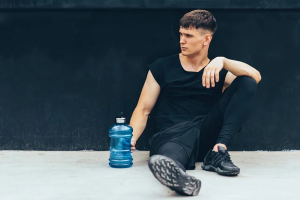 Bonito jovem descansando após o treino, com uma garrafa de água no fundo preto. Saudável masculino fazendo uma pausa depois de se exercitar ao ar livre . — Fotografia de Stock