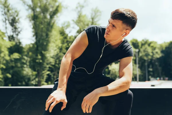 Retrato horizontal do jovem descansando após o exercício, ouvindo a música de fones de ouvido ao ar livre. Jovem descanso masculino muscular após um treino duro no sportsground. Pessoas e conceito de desporto . — Fotografia de Stock