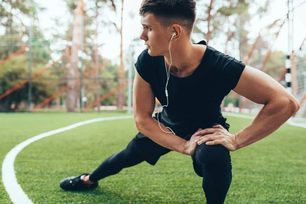 Outdoors image of muscular young man streching out against nature background. Athlete man looking away, stretching his leg and listening the music on earphones. Sport, lifestyle and people concept. — Stock Photo, Image