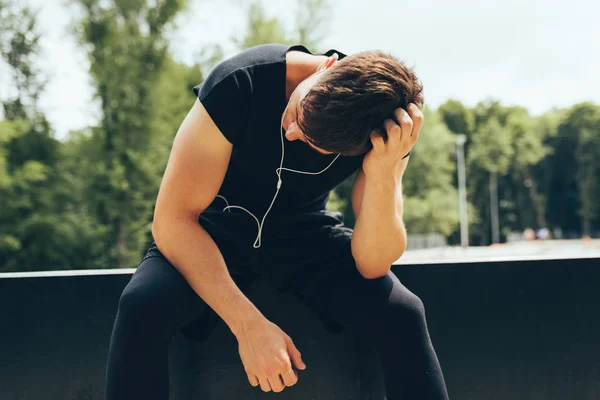 Hombre de fitness cansado descansando después de correr, escuchando la música de los auriculares al aire libre. Joven descanso masculino muscular después de un duro entrenamiento en el campo de deportes. Concepto de personas y deporte . —  Fotos de Stock