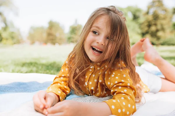 Sonriente niña linda acostada en la manta en la hierba verde, hablando con sus padres. Adorable niño divirtiéndose al aire libre durante el picninc con la familia en el parque. Concepto de infancia feliz . — Foto de Stock