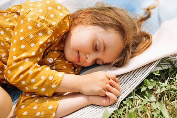 Κοντινό πορτρέτο από ένα χαριτωμένο κοριτσάκι που κείτεται στην κουβέρτα στο πράσινο γρασίδι και κοιμάται. Αξιολάτρευτο παιδί χαλαρωτικό, έχοντας χρόνο ύπνου σε εξωτερικούς χώρους κατά τη διάρκεια του picninc με την οικογένεια στο πάρκο. Έννοια της παιδικής ηλικίας. — Φωτογραφία Αρχείου