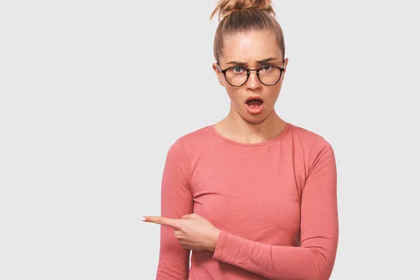 Incroyable et excitée jeune femme portant une tenue décontractée et des lunettes transparentes, la bouche ouverte en signe de panique attire votre attention sur l'espace de copie vierge. Femme perplexe posant sur un mur de studio blanc — Photo