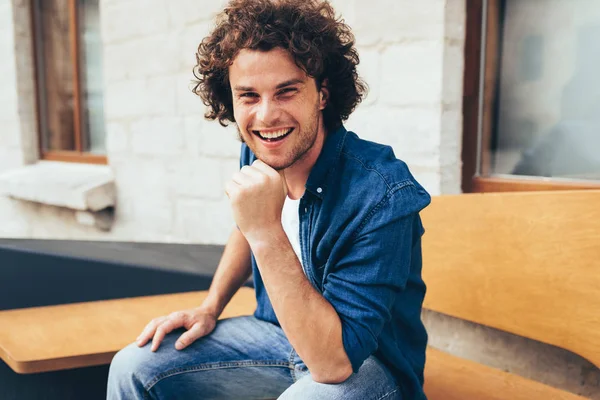 Sorrindo jovem bonito inteligente com cabelo encaracolado, olhando para a câmera e sentado no banco na rua da cidade. Estudante do sexo masculino feliz relaxar ao ar livre. Pessoas e conceito de estilo de vida — Fotografia de Stock