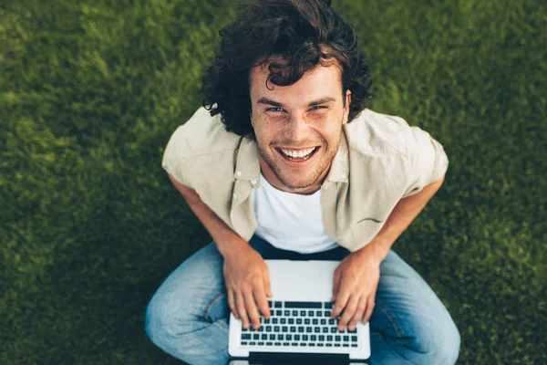 Pohled na šťastného studenta, který sedí na zemi a pracuje na laptopu. Usměvavý muž s kudrnatými vlasy, který používá přenosný počítač k konverzaci online s přítelem, na trávě. Mužské psaní na počítač v parku — Stock fotografie