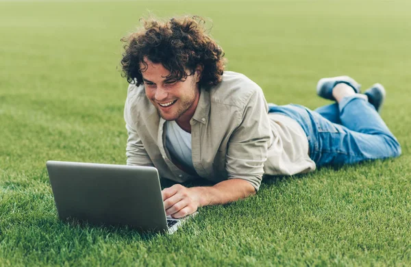 Veselý mladý muž na volné noze, který sedí na zemi a pracuje na laptopu. Usměvavý muž s kudrnatými vlasy, který používá přenosný počítač k konverzaci online s přáteli, na trávě. Mužské psaní na počítač v parku — Stock fotografie