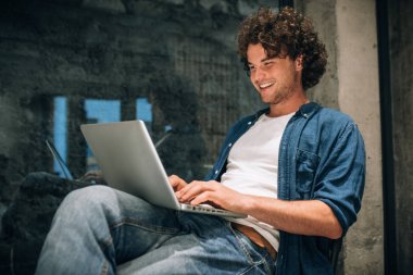 Online çalışma için dizüstü bilgisayar kullanarak kıvırcık saçlı mutlu genç adam Portresi. Akıllı serbest adam müşterileri ile online sohbet. Yakışıklı erkek haber okuyor, ofiste ki dizüstü bilgisayarına yazıyor