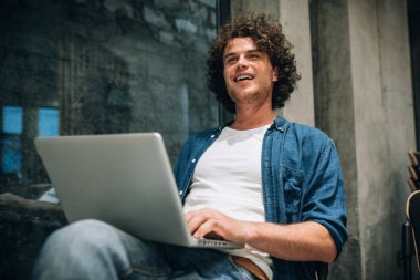 Kıvırcık saçlı yakışıklı genç adam online çalışmak için dizüstü bilgisayar kullanıyor. Akıllı serbest adam müşterileri ile online sohbet. Akıllı erkek okuma haber, modern ofiste onun laptop bilgisayarda yazarak