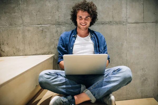 Slimme jonge man met krullend haar met behulp van laptop voor online werken, surfen op het Internet, zittend op de trap aan betonnen muur. Knappe mannelijke lezen en typen op zijn laptop computer in het kantoor — Stockfoto