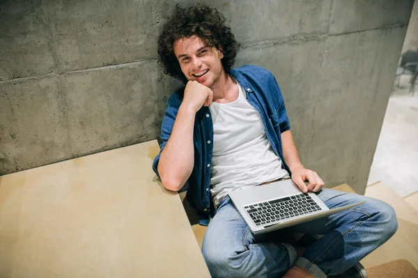 Estudante alegre com cabelo encaracolado usando laptop para aprender on-line, navegando na Internet, sentado nas escadas na parede de concreto. Sorrindo leitura masculina e digitação em seu computador portátil no escritório — Fotografia de Stock