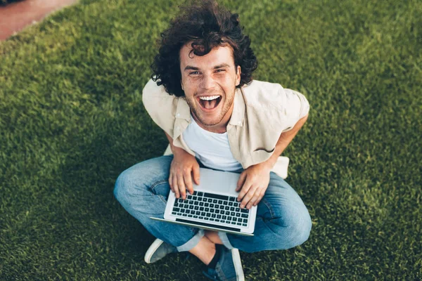 Gelukkige man zittend op de grond in het Park, werken op laptop. Lachende man met krullend haar met behulp van laptop voor online chatten met vrienden, zittend op het gras buiten. Technologie, Business, mensen — Stockfoto