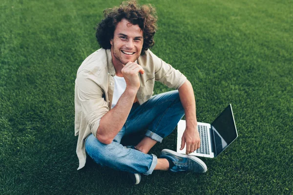 Candid shot van vrolijke freelance man met krullend haar met behulp van laptop om buitenshuis te werken, verbonden met gratis draadloos, zittend op het groene gras. Knappe mannelijke typen op zijn laptop computer in het Park — Stockfoto