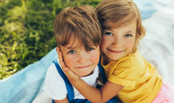 Nahaufnahme Porträt lächelnder zwei Kinder, die auf der Decke im Freien spielen. Schwester umarmt ihren kleinen Bruder im Park. Kinder haben Spaß an der Sonne. — Stockfoto