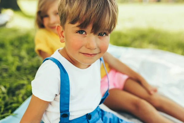 Закройте портрет улыбающегося мальчика, играющего на одеяле. Милый маленький мальчик отдыхает в парке с сестрой, проводящей время вместе. Очаровательные дети веселятся на солнце . — стоковое фото