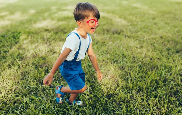 Immagine orizzontale di bambino felice che indossa pantaloncini blu e bicchieri di carta rossi che giocano sullo sfondo della natura. Bambino allegro che corre sull'erba verde nel parco. Ragazzo divertirsi alla luce del sole all'aperto — Foto Stock