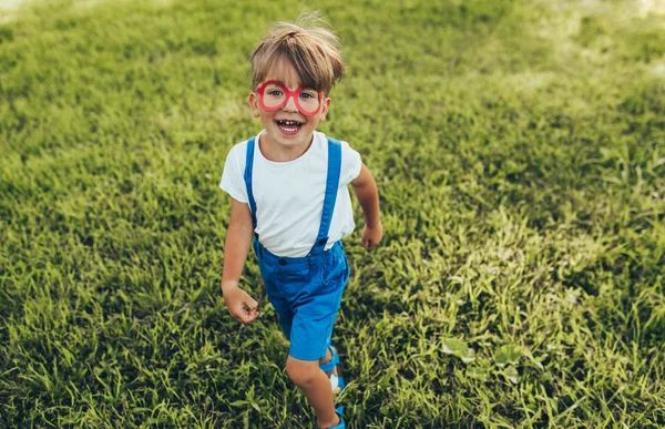 All'aperto immagine di felice bambino che indossa pantaloncini blu, t-shirt bianca e bicchieri di carta rossa che giocano sullo sfondo della natura. Bambino allegro che corre sull'erba verde nel parco. Ragazzo si diverte . — Foto Stock