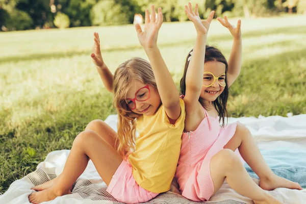 Lächelnde lustige Kinder, die Papierbrillen mit erhobenen Händen tragen, die Geburtstagsparty im Freien feiern und auf der Decke sitzen. zwei kleine Mädchen haben Spaß im Park. Freunde haben Spaß auf grünem Gras. — Stockfoto