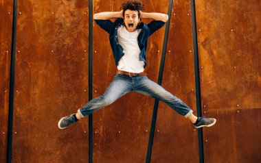 Kıvırcık saçlı yakışıklı genç adamın tam uzunlukta görüntü, gülümseyen ve modern binanın duvarında açık havada atlama. Mutlu akıllı öğrenci erkek kazanmak için çığlık ans atlama. İnsanlar ve duygular kavramı