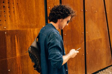 Avrupalı adamın açık havada durup cep telefonuna mesaj yazarak arkadan görüntü. Kıvırcık saçlı genç erkek, modern binanın duvarında cep telefonuyla gezinerek şehirde yürüyen güneş gözlüğü takıyor.