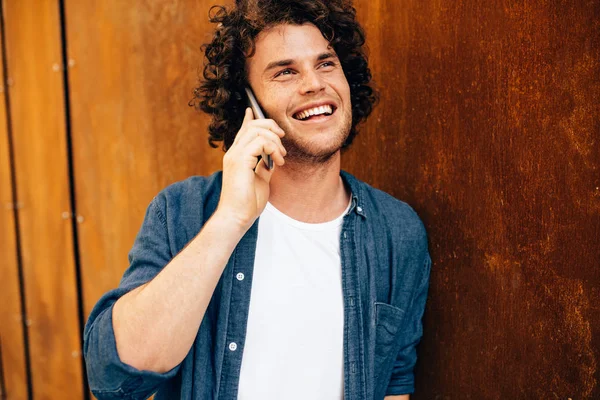 Retrato al aire libre de un joven feliz parado junto a un edificio moderno y hablando por teléfono móvil. Joven estudiante masculino con el pelo rizado haciendo una llamada en su teléfono celular en el fondo industrial . — Foto de Stock