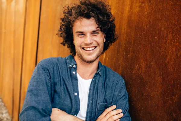 Retrato de belo jovem posando para propaganda, sorrindo e olhando para a câmera, em pé na parede do edifício ao ar livre. Retrato de estudante inteligente feliz do sexo masculino. Pessoas e conceito de emoção — Fotografia de Stock