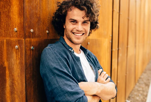 Jovem positivo com cabelo encaracolado, sorrindo e olhando para a câmera, em pé na parede do edifício de metal moderno ao ar livre. Retrato de estudante inteligente feliz do sexo masculino. Pessoas e conceito de emoção — Fotografia de Stock