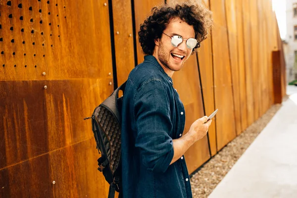 Achteraanzicht van gelukkige man buiten staan, het typen van berichten op de mobiele telefoon. Jonge man met krullend haar draagt zonnebril wandelen in de stad browsen op zijn mobiele telefoon op moderne industriële muur. — Stockfoto