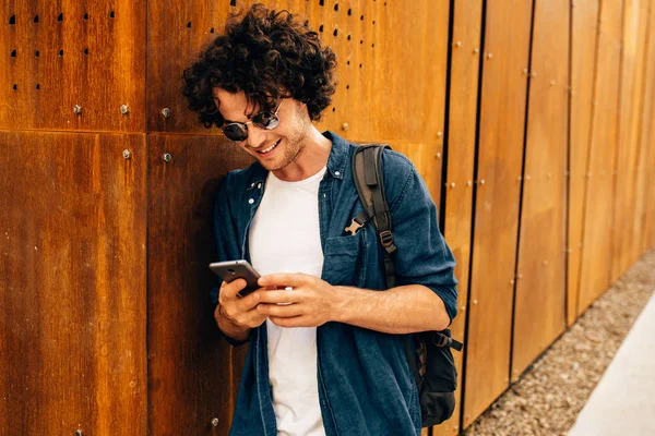 Close-up portret van knappe man staande buitenshuis, het typen van berichten op mobiele telefoon. Jonge man met krullend haar draagt zonnebril rusten buiten in de stad browsen op zijn mobiele telefoon op moderne muur — Stockfoto