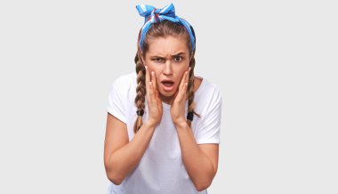 Beyaz t-shirt giyen şok güzel genç kadın Closeup portre ve ağız ile mavi kafa bandı beyaz arka plan üzerinde poz açtı. Şaşırmış kadın yanaklarını elleriyle tutar