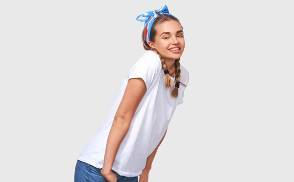 Очаровательная молодая женщина в белой футболке, голубой повязке с руками в карманах, улыбающаяся и смотрящая в камеру, изолированная на белом фоне студии — стоковое фото