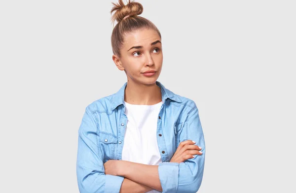 Mujer joven insatisfecha con las manos cruzadas, con atuendo casual con moño de pelo. Estudiante infeliz posando sobre la pared blanca del estudio. Concepto de personas y emociones —  Fotos de Stock