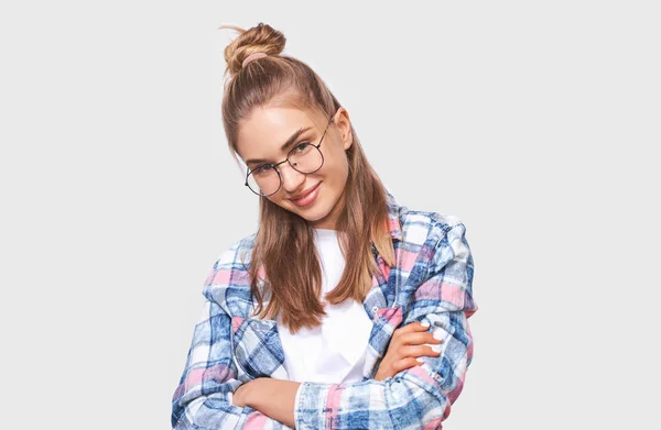 Positive young woman dressed in casual outfit, wearing round transparent eyewear with pleasant smile, keeps both arms crossed, looking to the camera and posing over white studio wall. People emotions — Stock Photo, Image