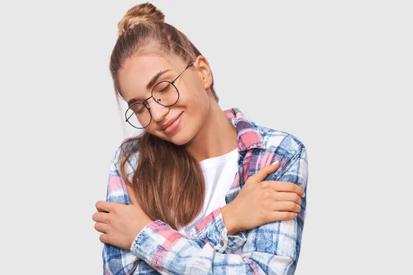 Jovem europeia satisfeita vestida com roupa casual, vestindo óculos redondos transparentes com sorriso agradável, com olhos fechados, posando sobre a parede branca do estúdio. Conceito de pessoas e emoções — Fotografia de Stock