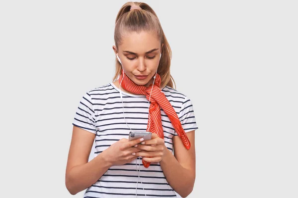 Displeased young woman using cell phone, messaging to her boyfriend. Unhappy female frowning her face and chatting with her boyfriend, looking at screen of smart phone, listening music in earphones. — Stock Photo, Image